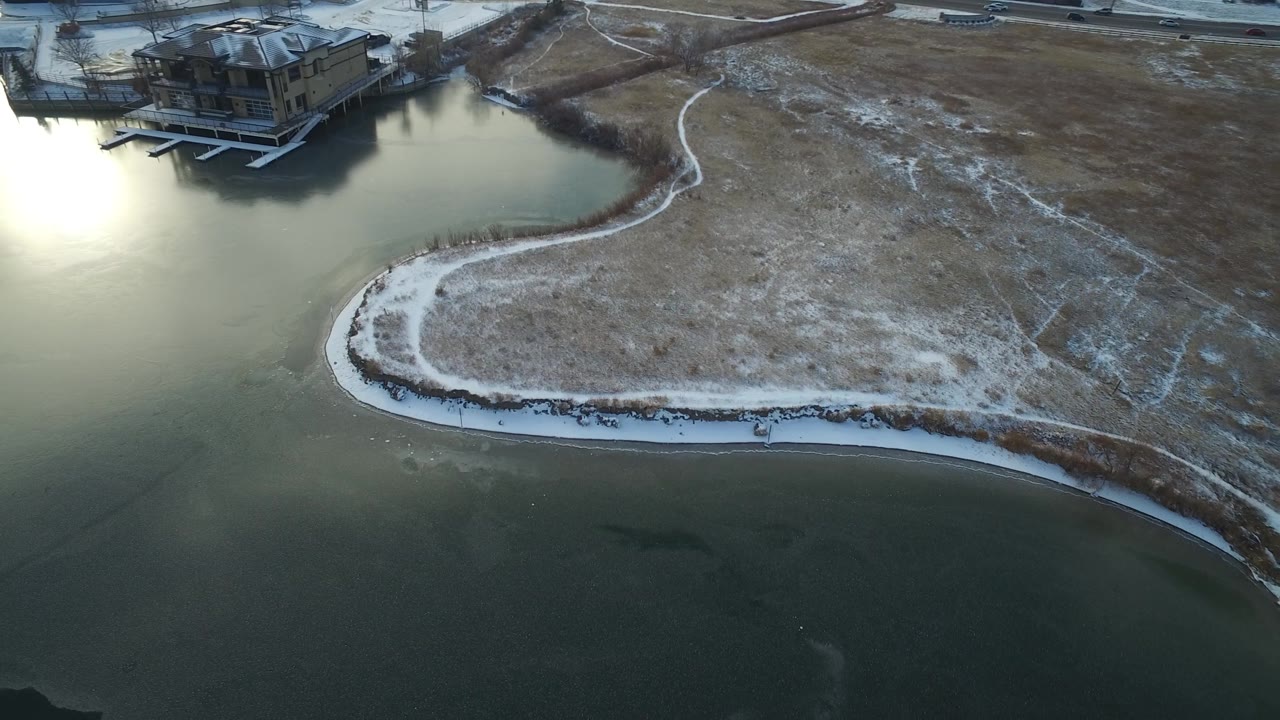 Aerial view over Hidden Lake Colorado with a bright reflection on the water, a snow-lined bend in the shore, scattered rocks, dry grass, and no visible people.