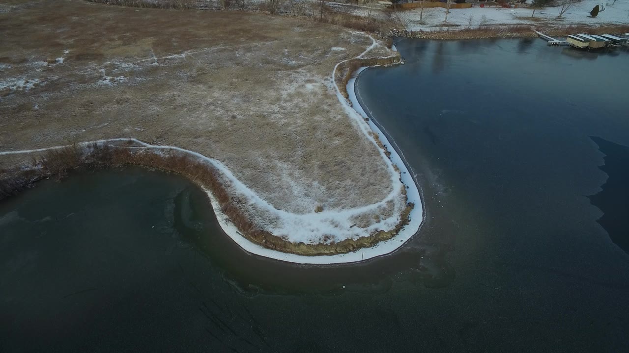 Aerial frame over Hidden Lake Colorado with the peninsula more centered, snow outlining the shoreline, dry grass across the middle ground, dark water around it, and no visible people.