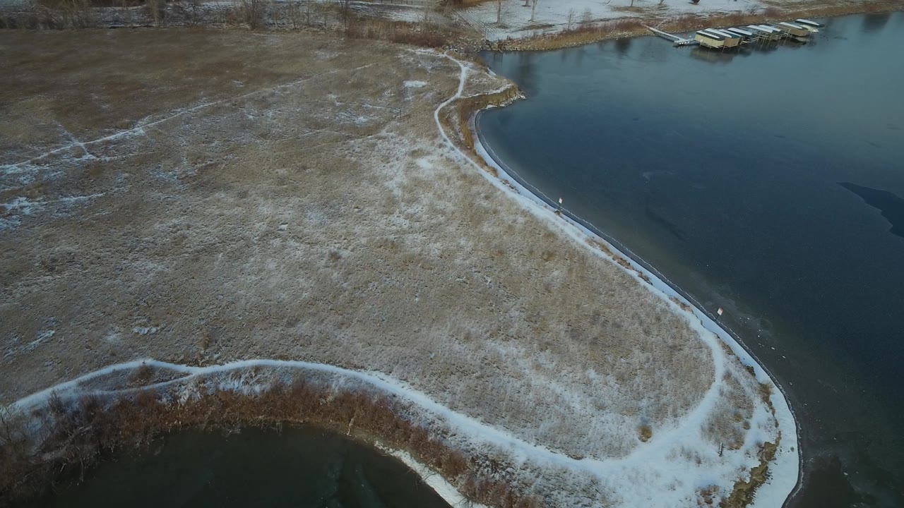 Aerial frame over Hidden Lake Colorado showing a curved snow-lined shoreline, brown winter grass, dark water, small docks near the top edge, and no visible people.