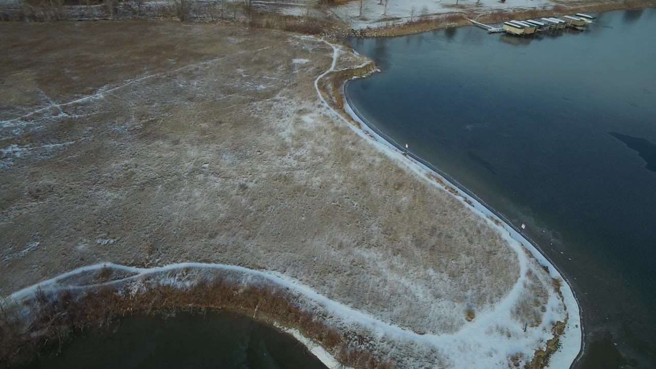 Hidden Lake Colorado appears from above with dark water on the right, pale snow outlining a narrow shore path, winter grass across the center, and no visible people.