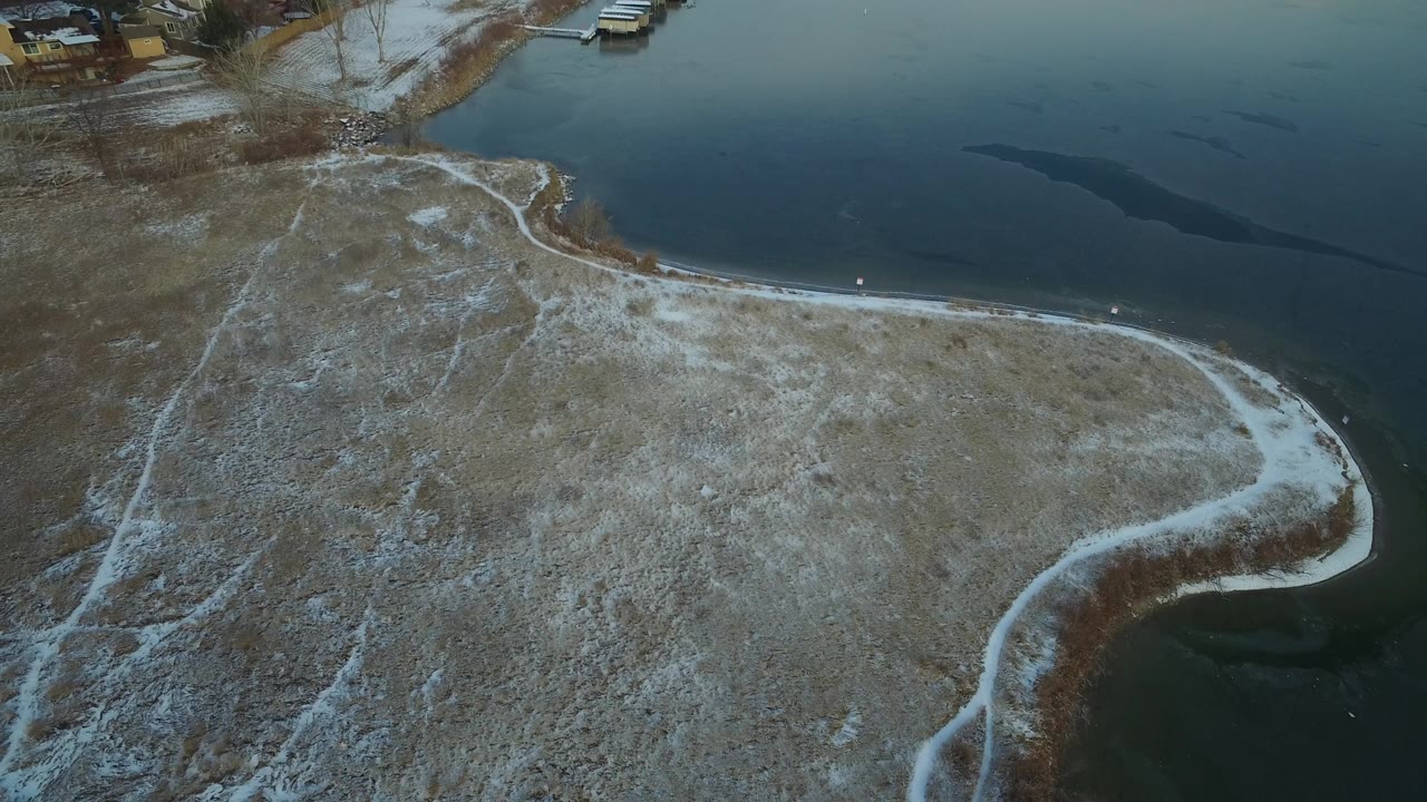 Aerial view in Hidden Lake, Colorado, with pale snow tracing paths across dry grass, a rounded shoreline at right, dark lake water above, and no visible people.