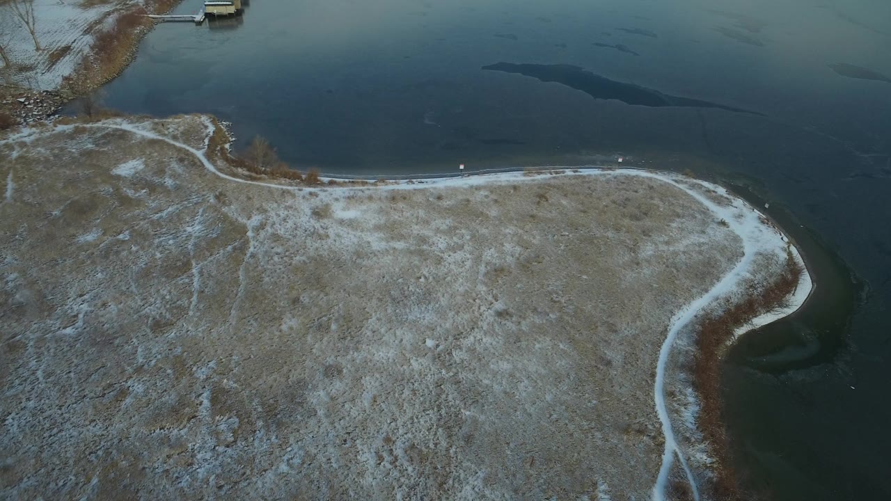 High aerial view over Hidden Lake, Colorado, showing a pale snow-dusted shoreline curving below dark green-blue water, with no visible people.