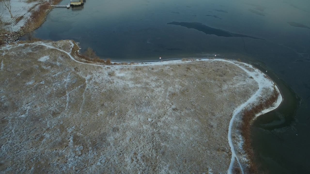 Birds-eye frame over Hidden Lake, Colorado, with the waterline running across the upper half and thin snow tracing the curved land edge, no visible people.