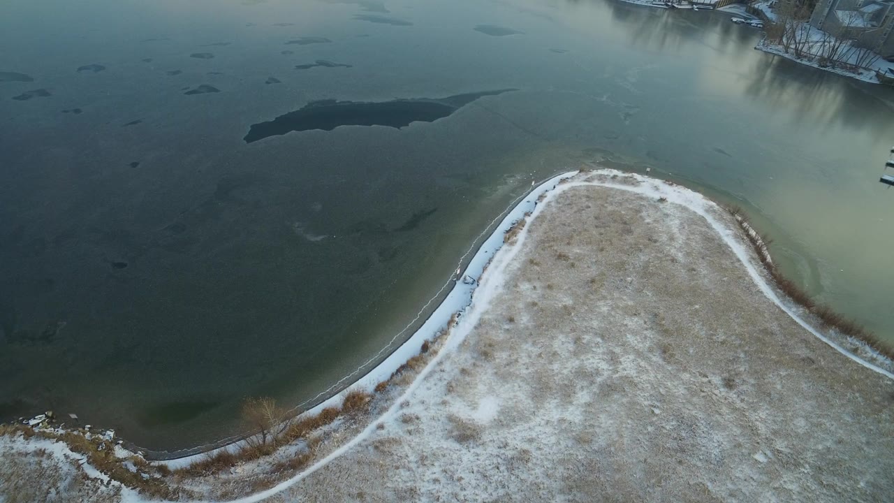 Overhead view at Hidden Lake, Colorado showing a snow-dusted curved shoreline, dark lake water, winter grass, distant docks, and no clearly visible people.