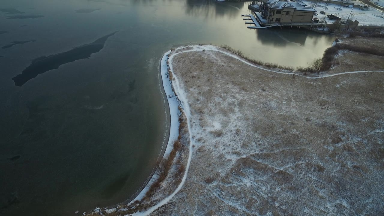 High aerial view over Hidden Lake, Colorado, with dark water, a curved snow-dusted shoreline, brown grass, docks, and no visible people.