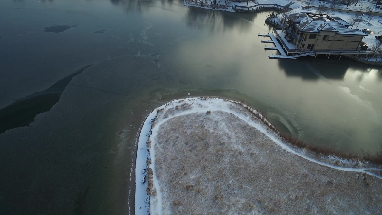 Hidden Lake, Colorado aerial still with the snow-covered curved bank lower in frame, muted frozen water across the center, waterfront houses near the top, and no visible people.