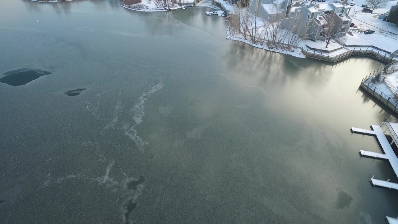 Aerial view over Hidden Lake, Colorado showing green-gray frozen water, dark ice openings, snowy banks, docks, private homes, and no visible people.