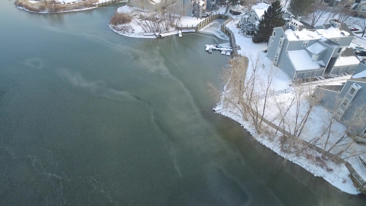 Overhead view of Hidden Lake, Colorado with green-blue icy water, a snow-covered shoreline, waterfront homes, docks, and no visible people at the opening moment.