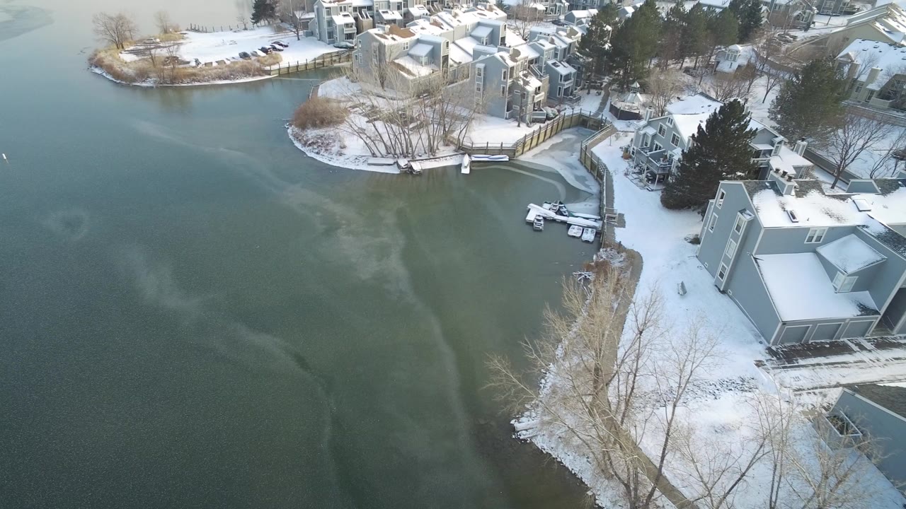 Elevated aerial frame over Hidden Lake, Colorado, showing green-gray frozen water, snow-covered residential shoreline, docks, small boats, bare trees, and no visible people.