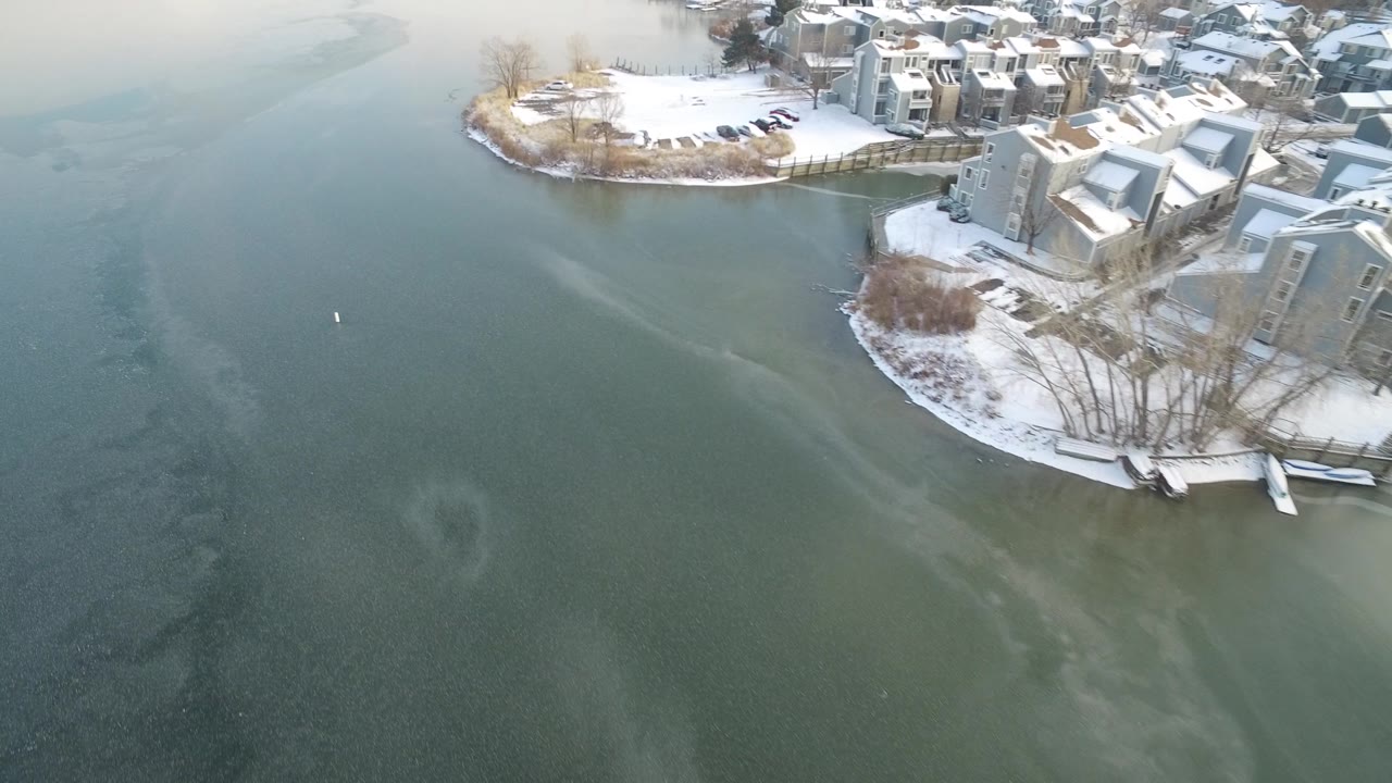 An aerial view at Hidden Lake, Colorado centers the textured lake surface with pale ice streaks below snowy homes and docks, with no visible people.