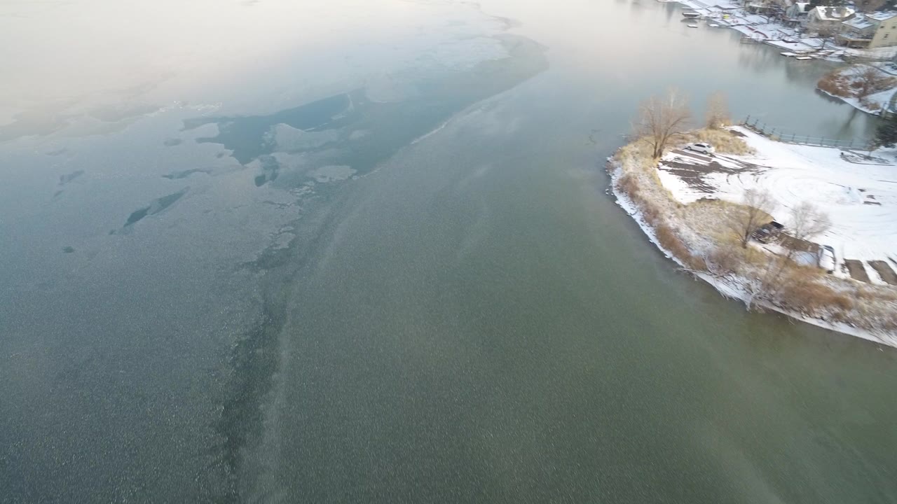 Aerial view of icy blue green lake water beside a snowy shoreline point.
