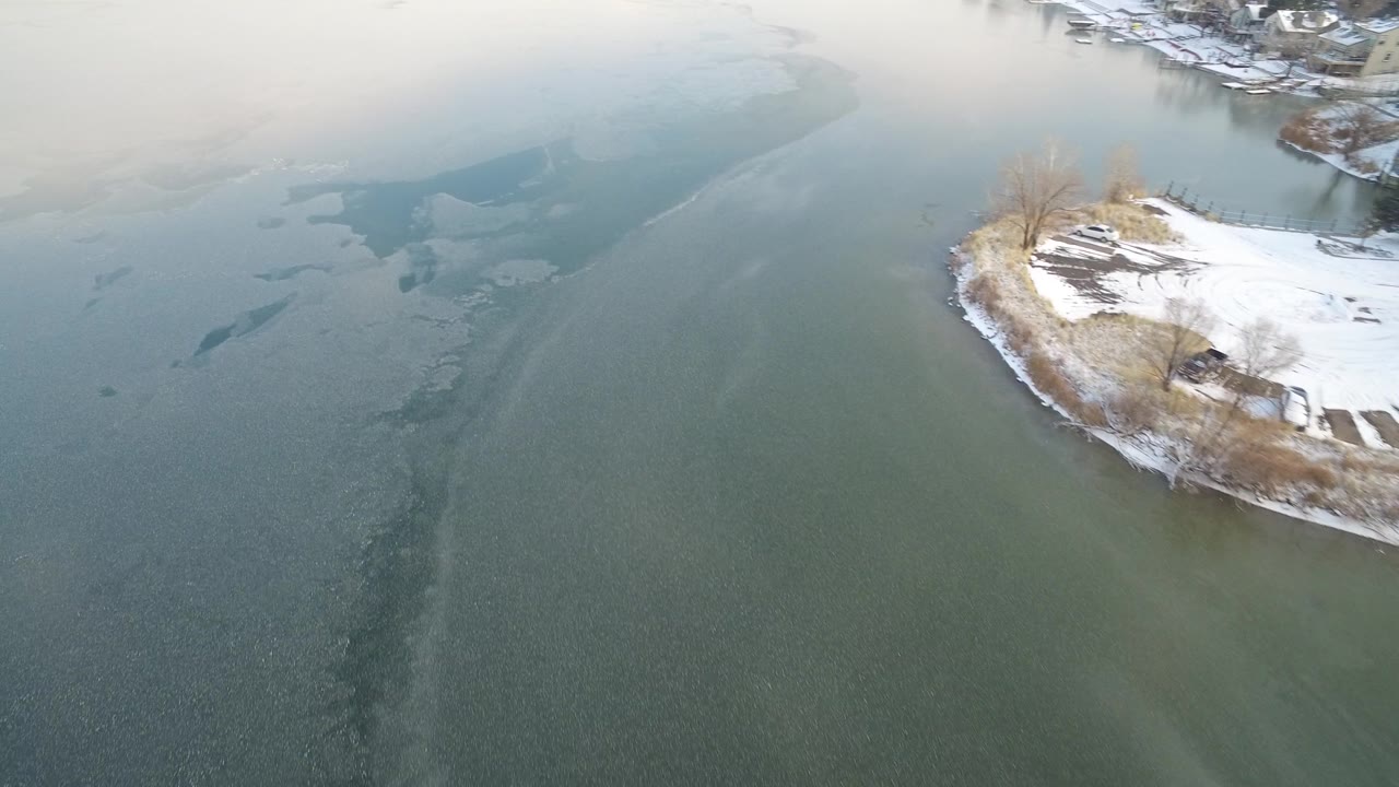 Aerial view of Hidden Lake, Colorado, with the shoreline point shifted farther right, pale ice bands across the water, distant shore edges, and no visible people.