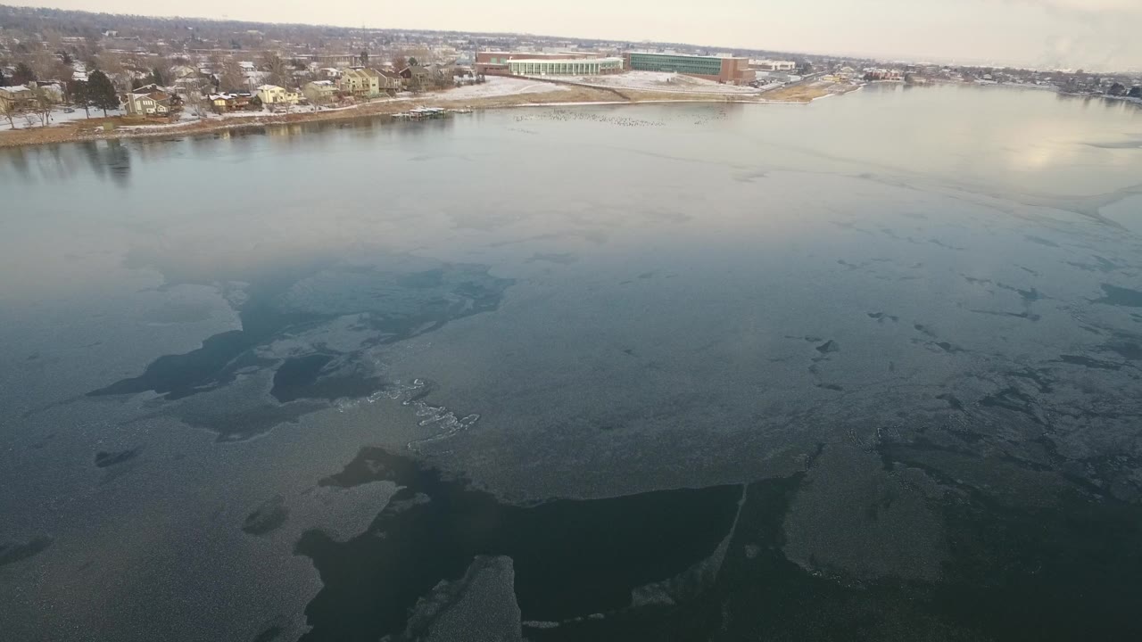Aerial view over a frozen lake beside a snowy residential shoreline.