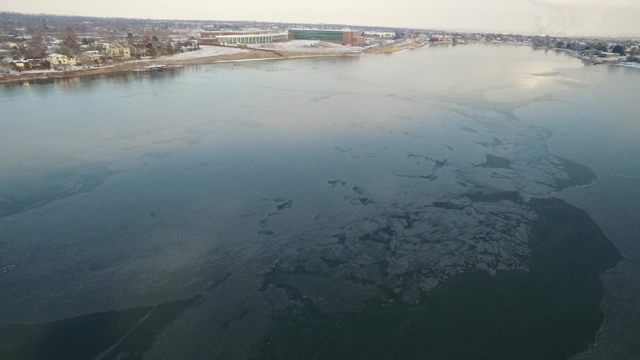 Early frame over Hidden Lake, Colorado shows a wide river with dark ice patches, a snowy shoreline, distant homes, a large low building, and no visible people.