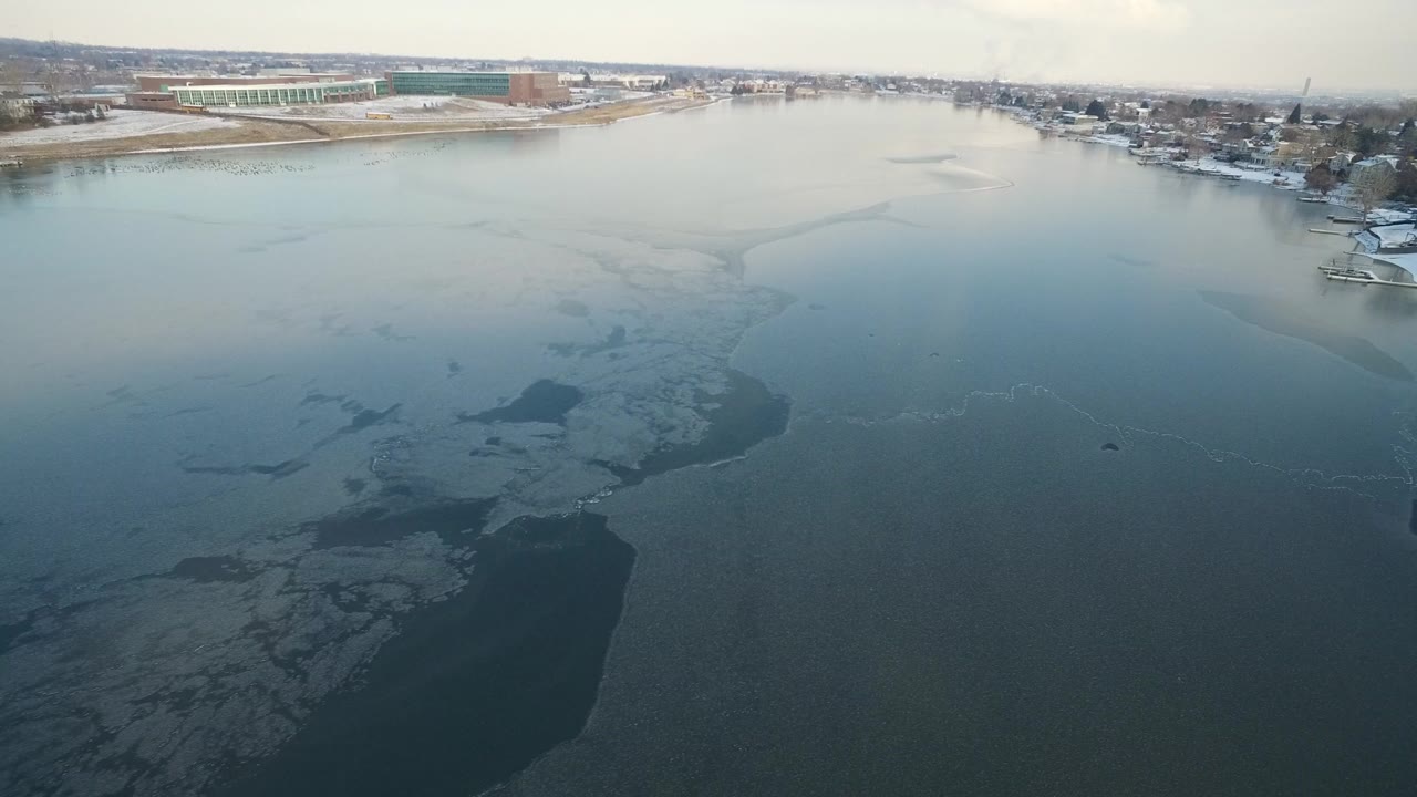 Aerial view over dark lake water with ice patches and distant shoreline buildings.