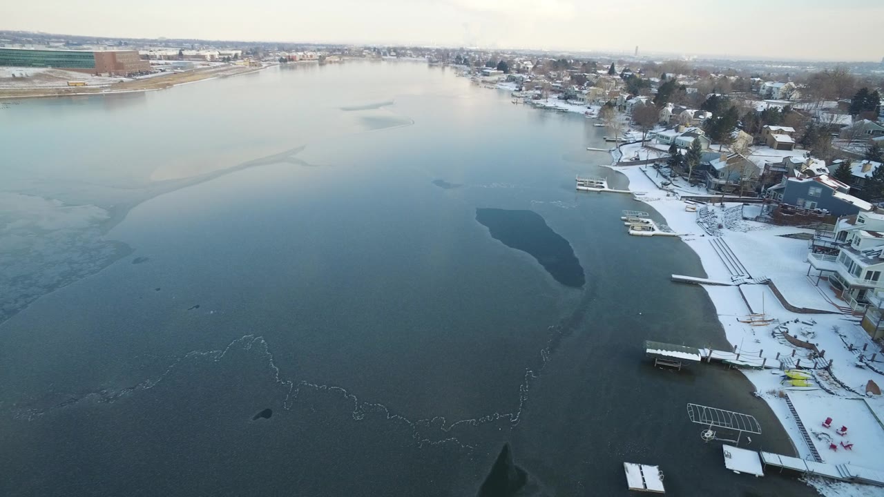 Aerial view over a partially frozen lake beside snowy waterfront homes.