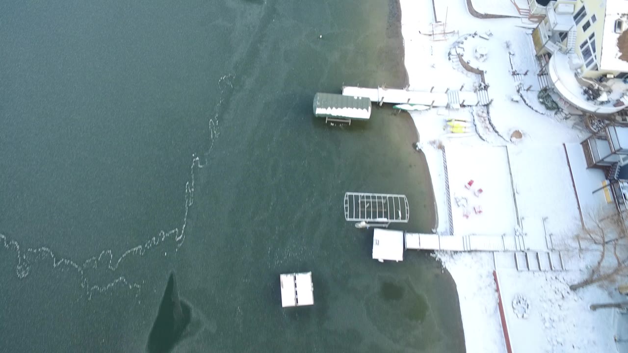 Overhead view of lake water, snowy shoreline, docks, and lakeside buildings.