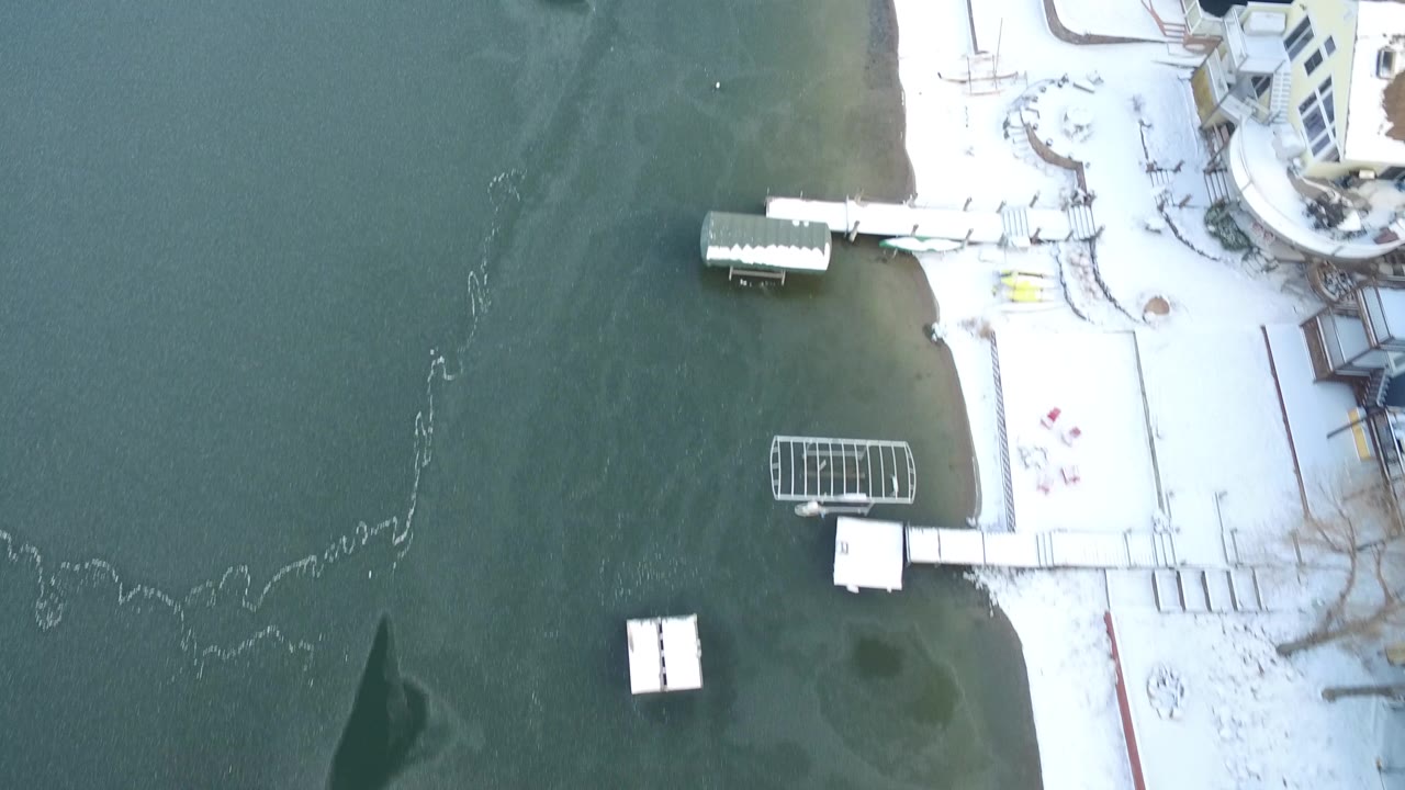 Aerial overhead frame from Hidden Lake, Colorado with lake water on the left, bright snow along the shore, small docks, walkways, and building edges, with no visible people.
