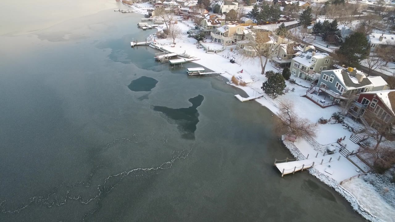 Hidden Lake, Colorado shoreline from above, with a larger open-water patch near center, snow on yards and roofs, and multiple docks extending into the icy lake.