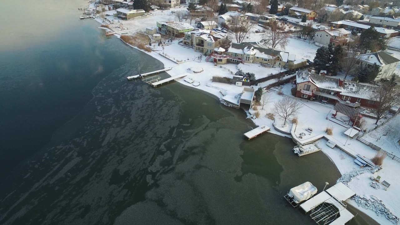 Aerial view over Hidden Lake, Colorado showing dark lake water, thin ice patterns, snow-covered yards, docks, and lakeside homes with no visible people.