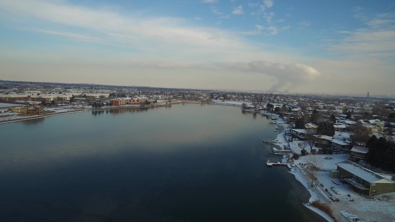 Elevated view over Hidden Lake, Colorado, with dark water in the foreground, snowy shorelines, low buildings across the bay, pale morning sky, and no visible people.