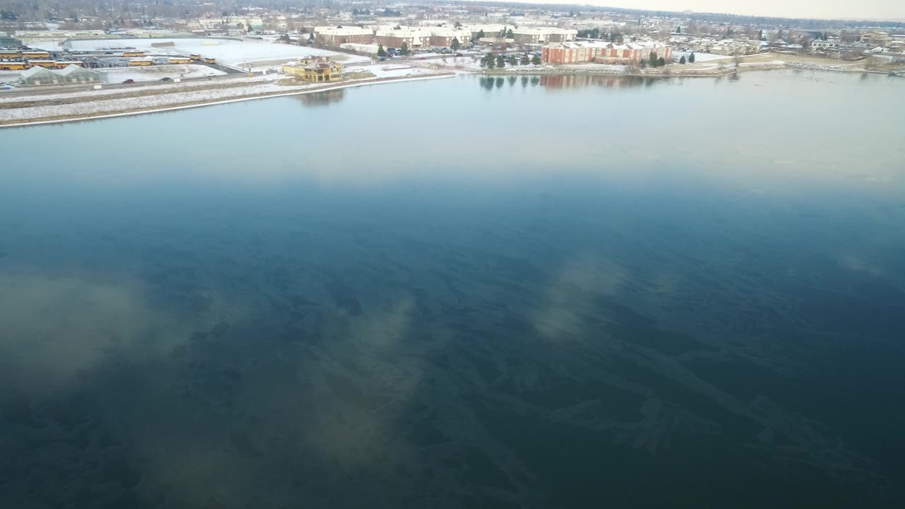 Aerial view over blue-green lake water toward a snowy residential shoreline.