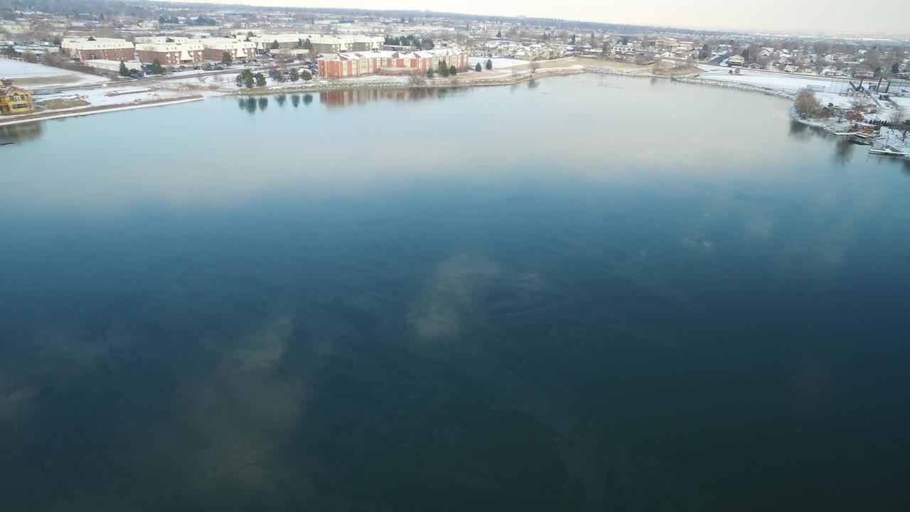 Aerial view over dark lake water with snowy shoreline and buildings beyond.