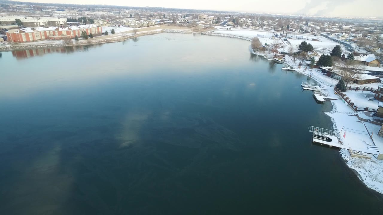 Aerial view over dark lake water toward a snowy shoreline and distant buildings.