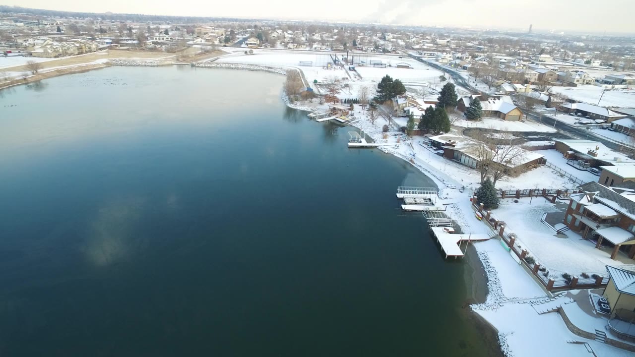 Aerial frame over Hidden Lake, Colorado showing dark lake water, a snow-covered residential shoreline, small docks, houses, leafless trees, and no visible people.