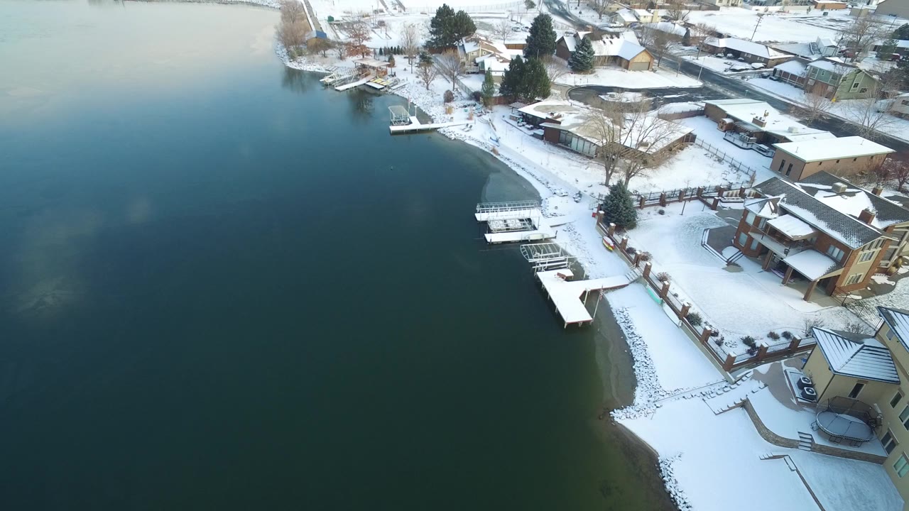 Aerial view of snow-covered lakeshore homes and docks beside dark green water.