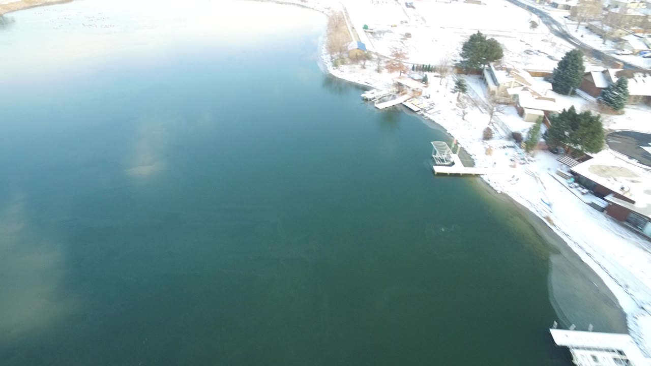 Aerial view of green-blue lake water beside a snowy shoreline with docks and trees.