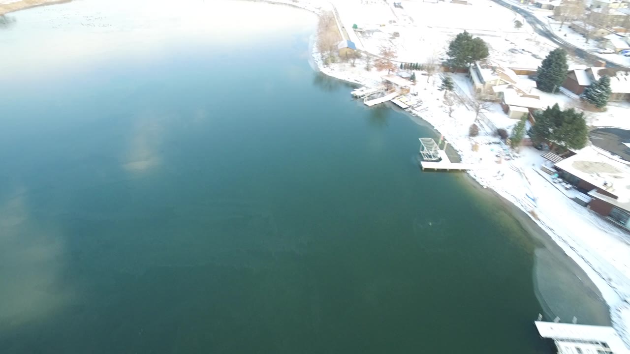 Aerial view in Hidden Lake, Colorado with broad green water filling most of the frame, snow on the right shoreline, clustered trees, docks, and no visible people.