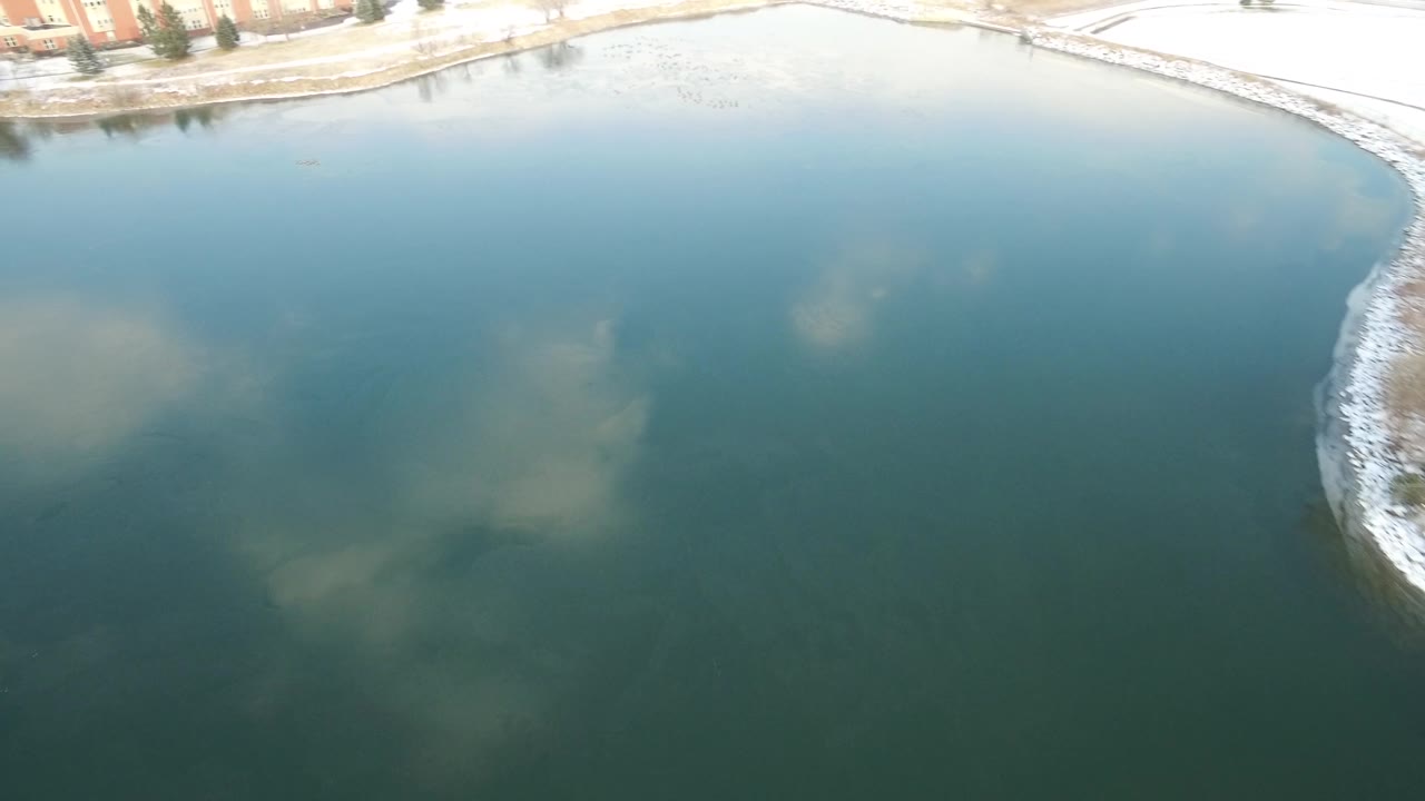 Aerial frame in Hidden Lake, Colorado with teal water filling most of the view, soft cloud reflections below, and a narrow shoreline with buildings along the top, with no visible people.