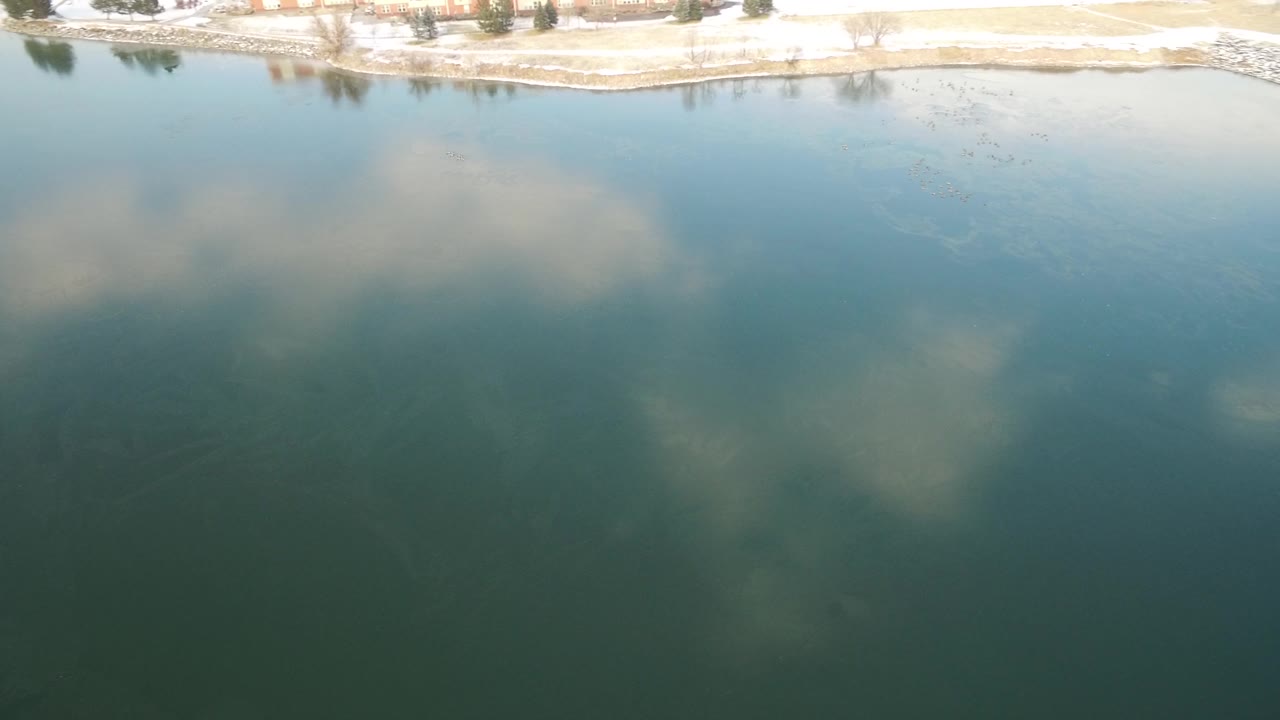 Aerial view over Hidden Lake, Colorado, with cloud reflections on blue green water, a snowy far shoreline, and no visible people.