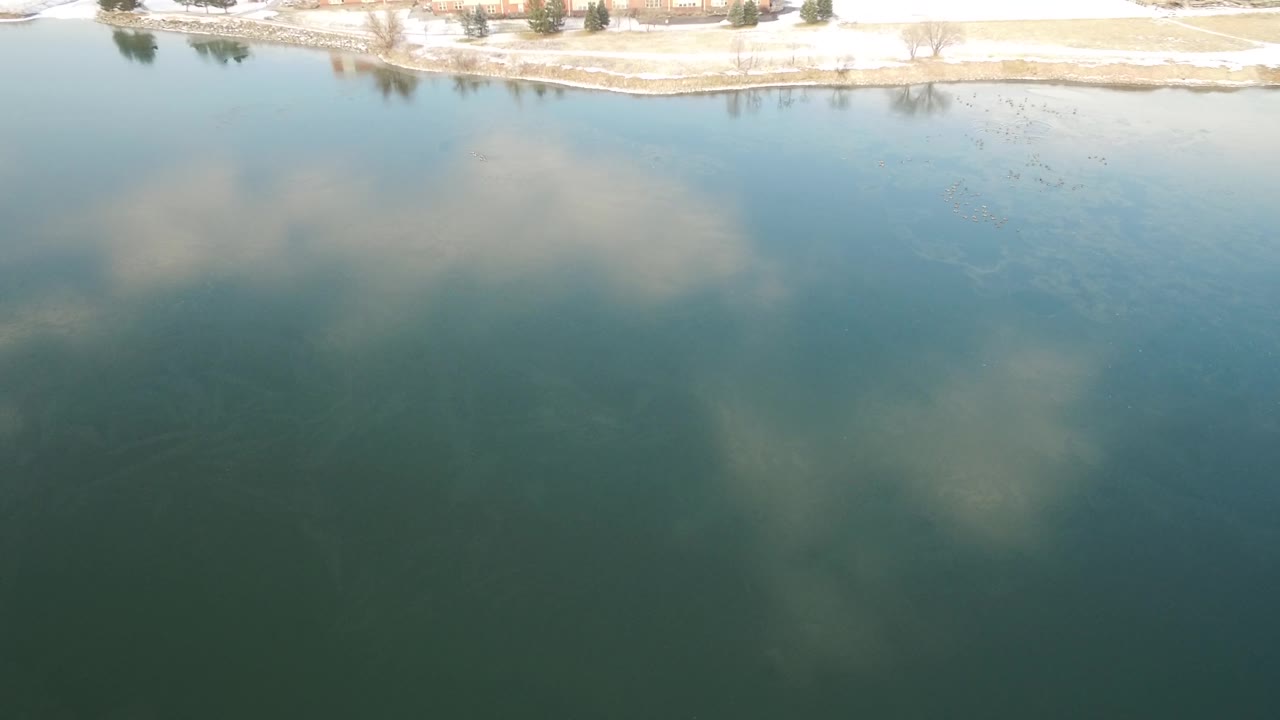 A wide aerial over Hidden Lake, Colorado, with muted cloud reflections, a winter shoreline, small buildings in the distance, and no visible people.