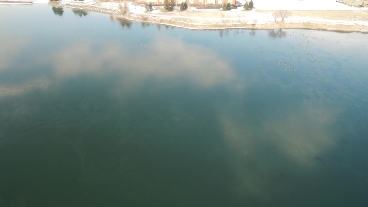 Aerial lake water with cloud reflection and snowy shore running across the upper frame.