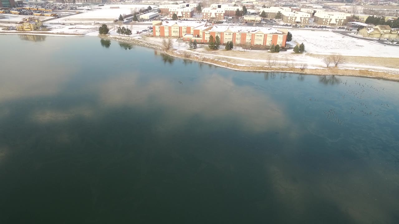 Aerial frame over Hidden Lake, Colorado showing blue-green water in the foreground, snow along the shore, and a large ivory and orange building beyond it, with no visible people.