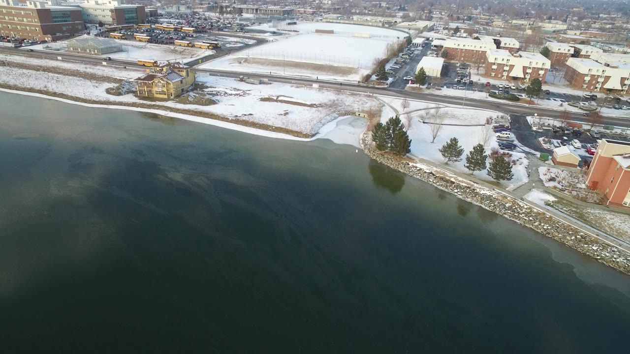 High aerial frame over Hidden Lake, Colorado, showing dark water, snow on the shoreline, roads, low buildings, and distant city blocks with no visible people.