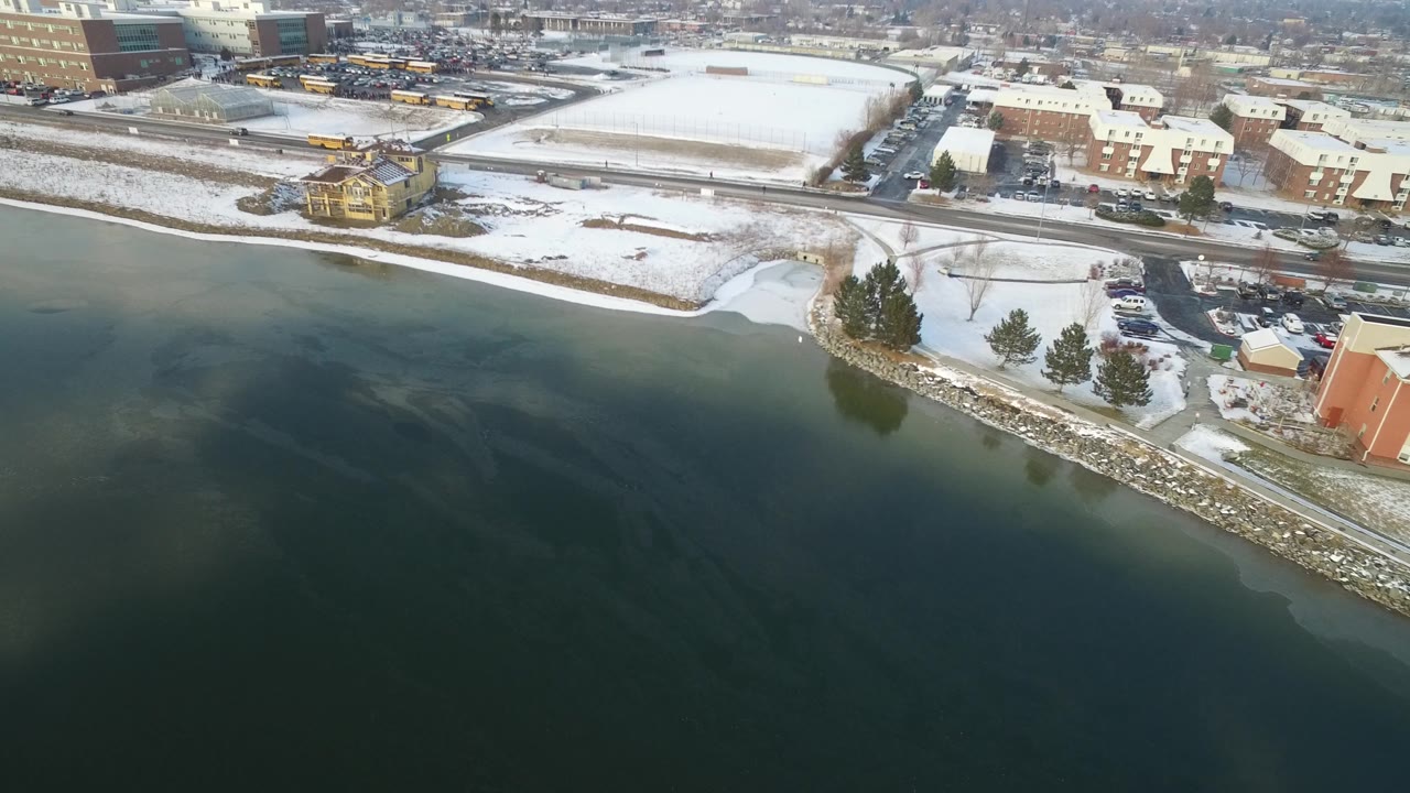 Hidden Lake, Colorado aerial frame with the lake occupying the lower half, a snowy bank and small inlet at the edge, and urban buildings beyond the road with no visible people.