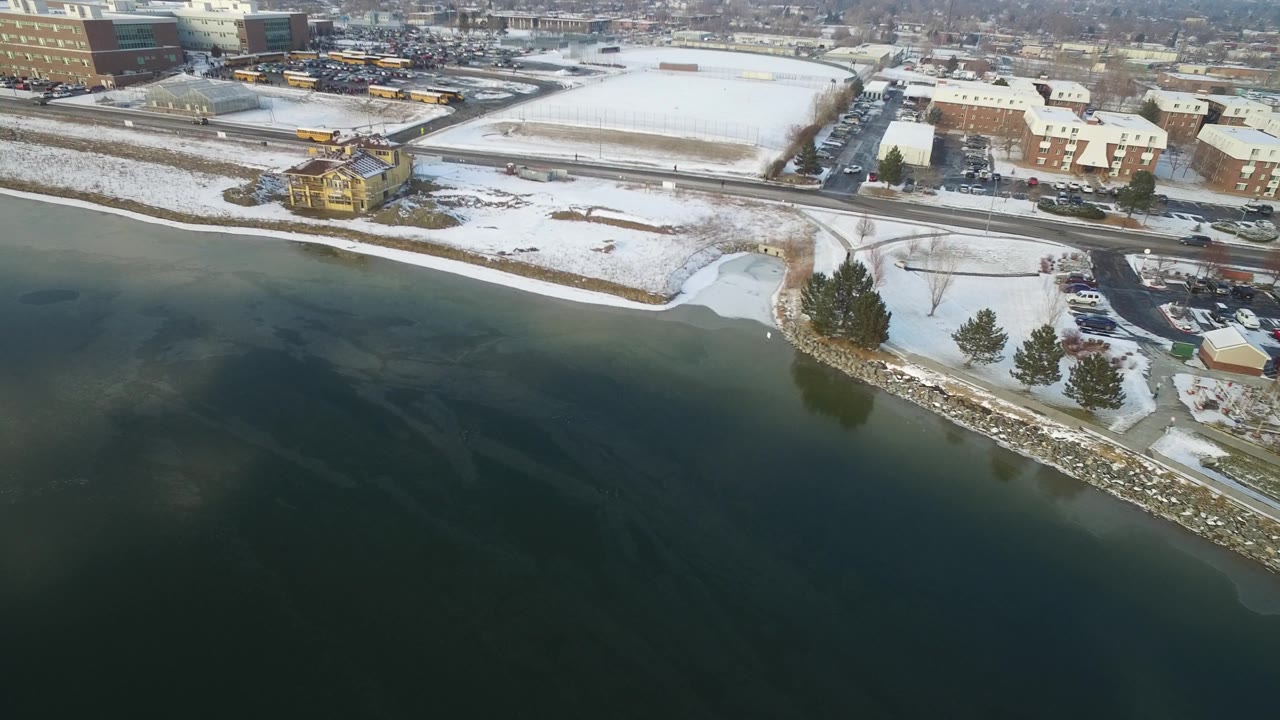 Aerial view of an icy lake edge, snowy lots, roads, buses, and city buildings.
