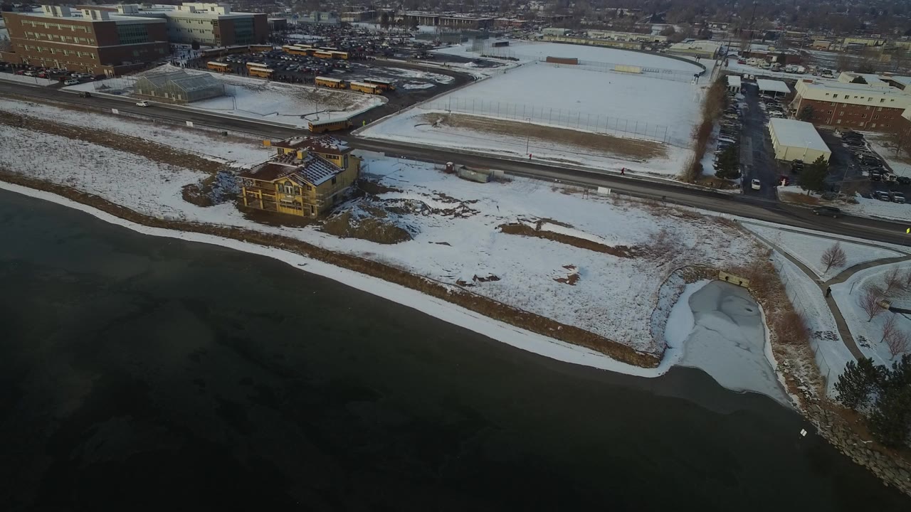 Aerial frame from Hidden Lake, Colorado showing a solitary house beside dark water, snow-covered ground, nearby roads, school buses in the distance, and no visible people.