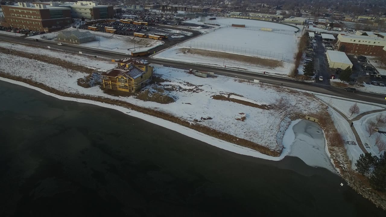 High aerial view in Hidden Lake, Colorado with the house near the lower shoreline, a road crossing behind it, snowy open lots, parked cars, yellow buses, and no visible people.