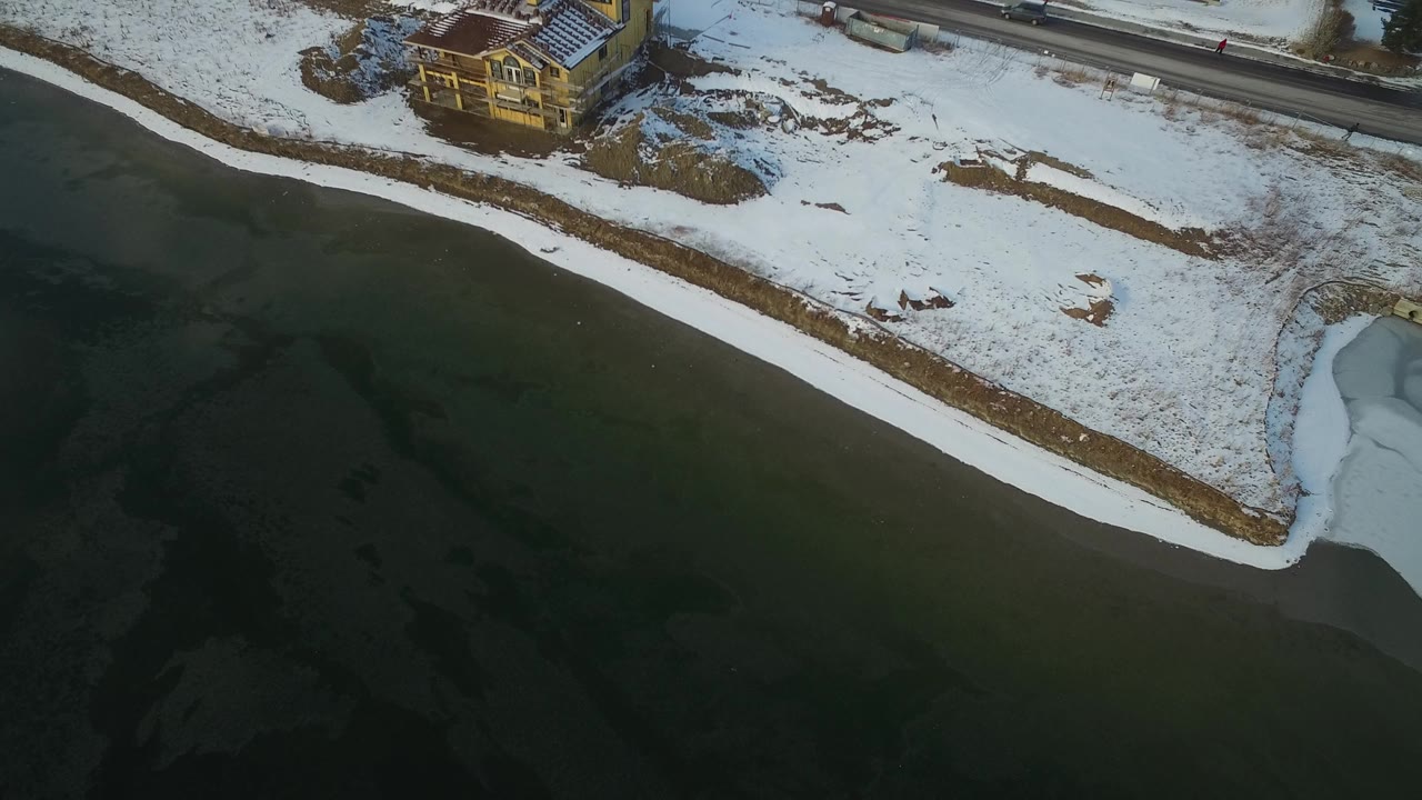 Overhead view of Hidden Lake, Colorado, with dark frozen lake water, a snow-covered shore, a small light-colored house near the top edge, and no visible people.