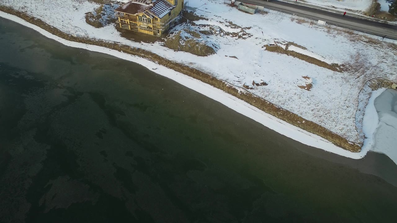 Snow-covered lakeside property and road viewed from above beside dark icy water.