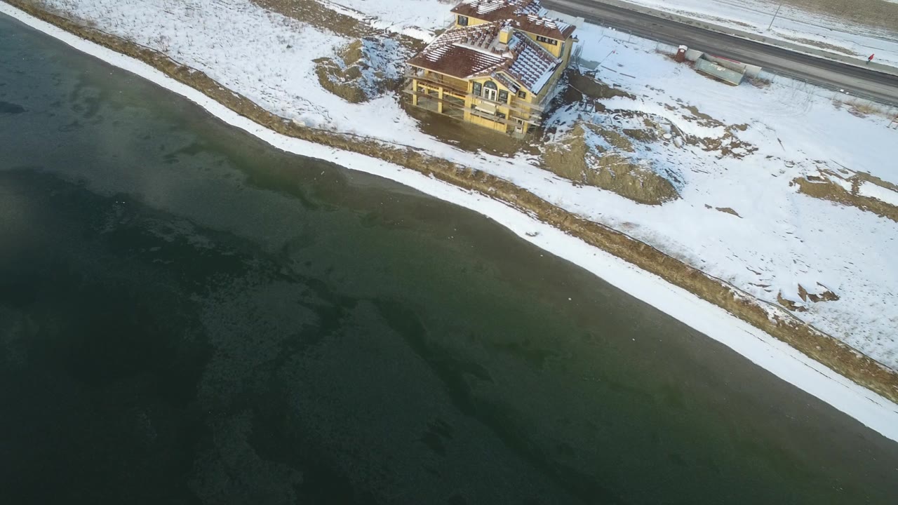 Aerial view of frozen water beside a snow-covered shoreline and lakeside house.
