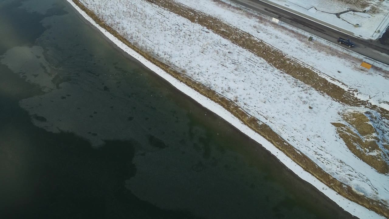 Overhead view of dark frozen lake, snowy shoreline, and road edge.