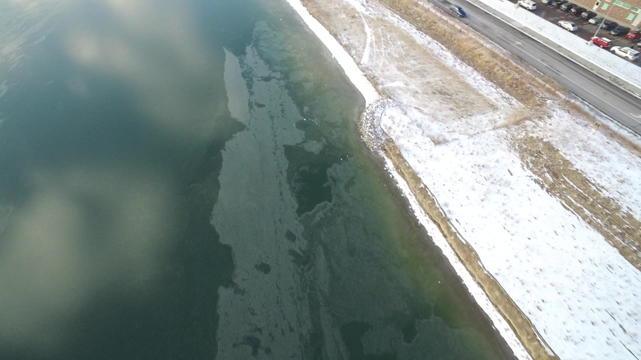 Aerial view of dark lake water beside a snowy shoreline and road.
