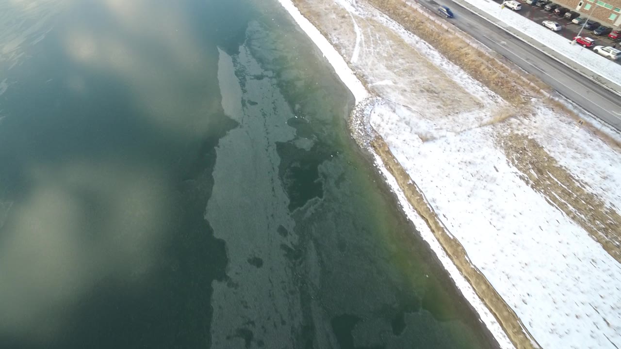 Aerial frame over Hidden Lake, Colorado, with the shoreline angled through the center, parked and moving cars near the road, cloud-like patches on the water, and no visible people.