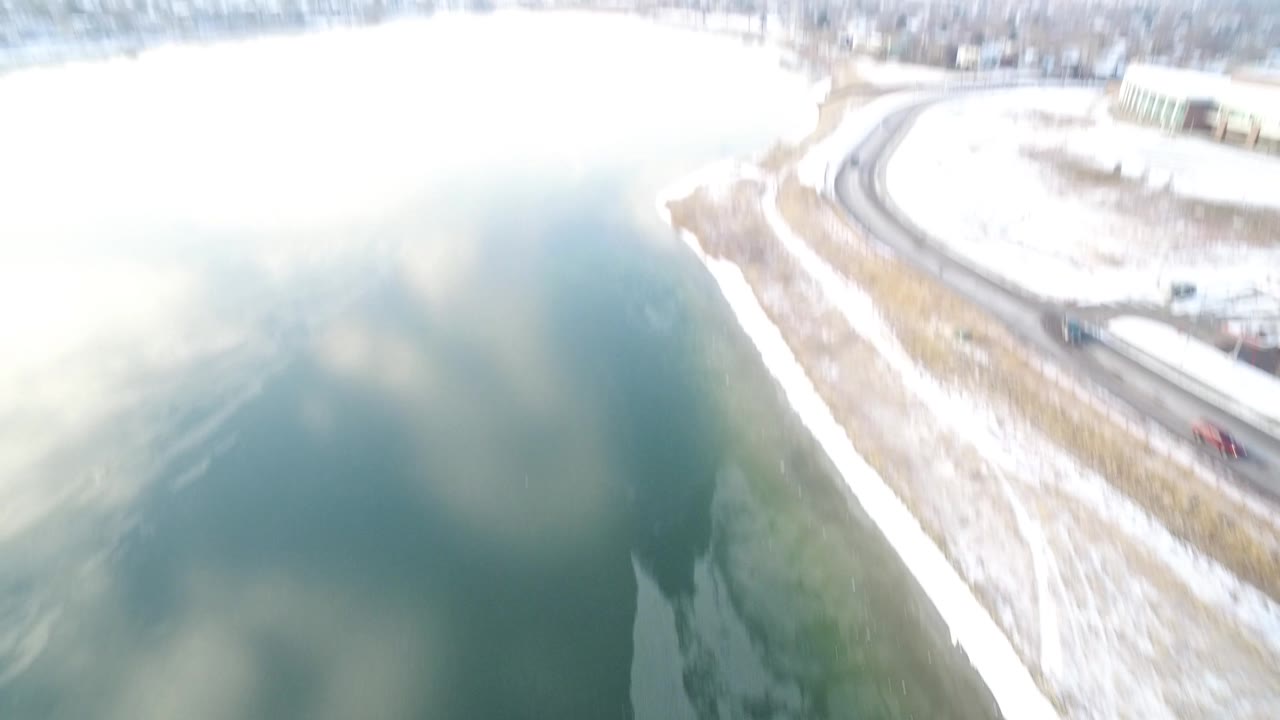 Drone-height view of Hidden Lake, Colorado with pale water, a snow-lined bank, a curving road, and no visible people.