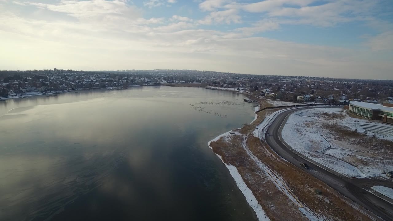 Aerial frame over Hidden Lake, Colorado showing calm water on the left, a curved shoreline road on the right, distant homes, light cloud cover, and no visible people.